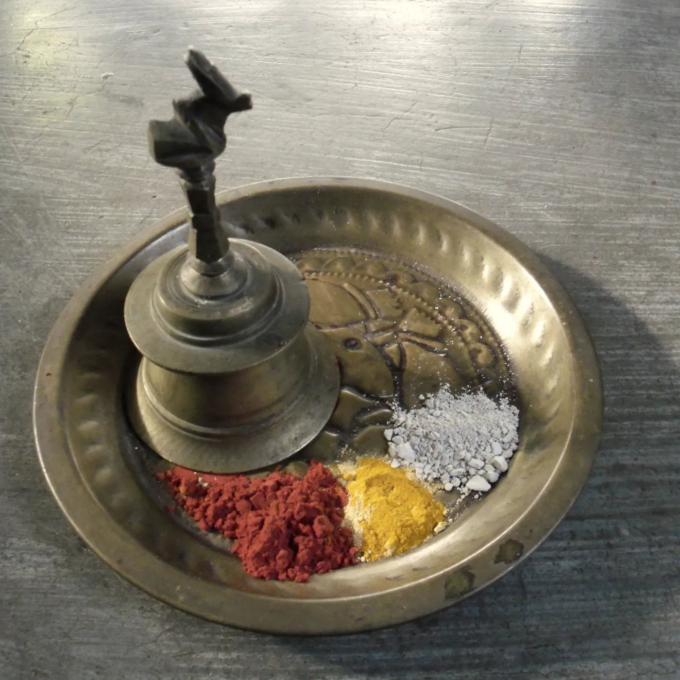 A brass puja plate holding a hand bell with kumkum, haldi and sacred ash